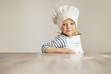 Laughing little girl as a chef in the kitchen in front of the empty table. Learn cooking.