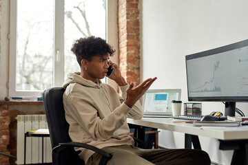 Irritated young male trader talking on the phone discussing stock market while looking at charts on computer