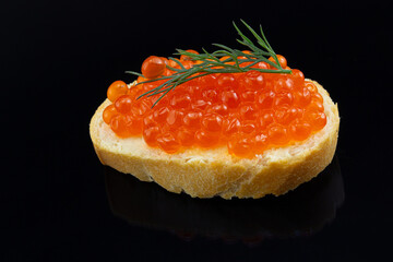 Close-up red caviar sandwich isolated on black background. Salted salmon caviar on wheat bread with fresh dill. Reflection in a shiny surface