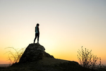 Silhouette of a woman hiker standing alone on big stone at sunset in mountains. Female tourist on high rock in evening nature. Tourism, traveling and healthy lifestyle concept.