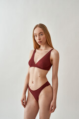 Attractive young slim caucasian woman with beautiful perfect body wearing burgundy underwear posing isolated over light gray background