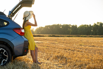 Young woman driver in yellow summer dress and straw hat standing near a blue car enjoying warm summer day at sunset.