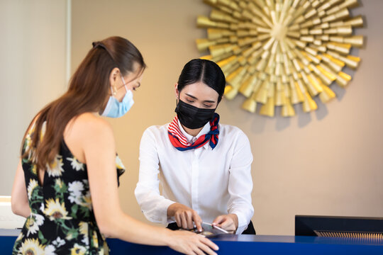 beautiful young asian woman receptionists working at a reception desk and holding key card or passport to customer. People wearing protective face mask prevent covid-19 virus - Powered by Adobe