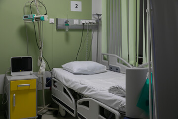 Hospital bed with a pillow in intensive care unit is surrounded by different medical devices: bedside patient monitor, oxygen delivery system, drip stand, infusion pump. Hospital environment is around