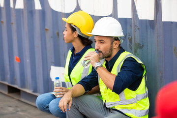 Tired hispanic male and female container worker in hardhat and uniform resting at container yard...