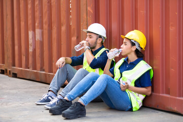 Tired hispanic male and female container worker in hardhat and uniform resting at container yard port of import and export. - obrazy, fototapety, plakaty