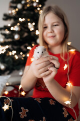 A teenage girl in a red festive T-shirt sitting with a hamster in her hands at the Christmas tree