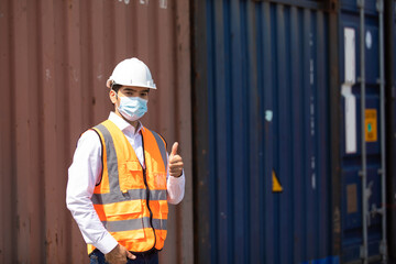 professional engineer man wearing protection face mask during coronavirus and flu outbreak and wearing safety hardhat helmet at container yard or cargo warehouse