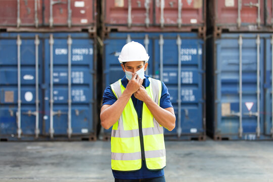 professional engineer man wearing protection face mask during coronavirus and flu outbreak and wearing safety hardhat helmet at container yard or cargo warehouse