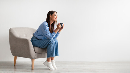 Full length of young woman having coffee break in cozy armchair against white studio wall, banner with free space © Prostock-studio