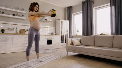 Curly pregnant sportswoman exercising with slam ball on fitness mat at home