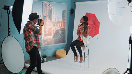 Stylish african model posing with red umbrella on bar high chair for fashion magazine photo shoot. Photographer taking photos on professional camera.