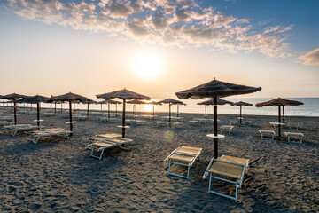 beautiful landscape of empty sunrise or sunset beach with chaise loungues and nice umbrellas with blue sea, sun glow and amazing cloudy sky on thr background