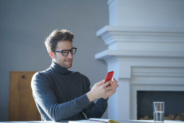 Young handsome german man in glasses sitting at table and using mobile phone at home