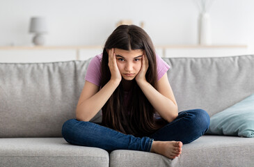 Fototapeta premium Upset Indian teenage girl sitting on couch, feeling bored, suffering from lack of communication, being trapped at home