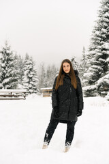 Fototapeta na wymiar Beautiful girl walking in a snowfall. Young woman in black puffer jacket posing in the winter forest.Outdoor portrait of pretty brunette lady. Winter concept