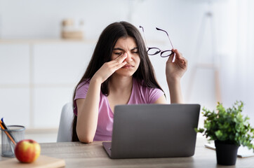 Indian teenage girl feeling tired, rubbing irritated eyes, sitting at desk with laptop, exhausted from online studies