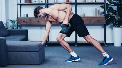 side view of shirtless sportsman training with kettlebell near grey sofa