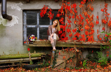 A beautiful young woman is seen relaxing by an old house.
She is surrounded by falling red autumn...