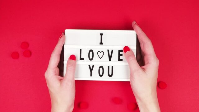 Hands of a young girl hold a white board with the inscription I love you on a red background with fluffy balls