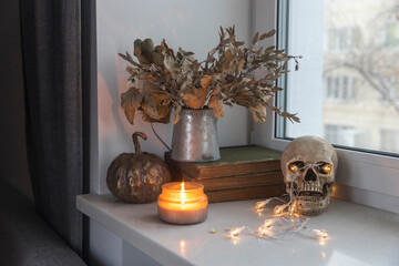 Bouquet of dried autumn leaves in a zinc jug, plastic skull, artificial gilded pumpkin, vintage books decorate the window for Halloween