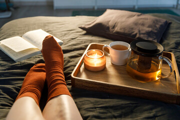 Soft photo of woman`s legs in the woolen socks on the bed with book and cup of tea and candle on the tray. Interior and home cosiness concept. Top view - obrazy, fototapety, plakaty
