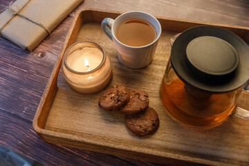 Interior and home cosiness concept. Top view. Burning candle in a jar, a cup of tea, a teapot with herbal tea and three oatmeal chocolate chip cookies on a wooden tray on the table