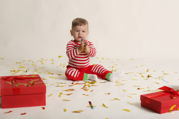 a little boy in a striped jacket and red pants sits on a white background with confetti and gift boxes