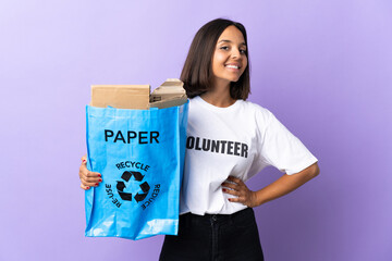 Fototapeta premium Young latin woman holding a recycling bag full of paper to recycle isolated on purple posing with arms at hip and smiling