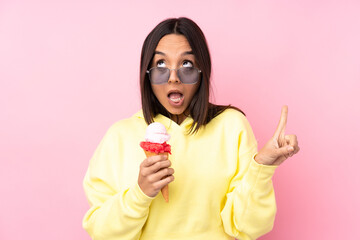 Young brunette girl holding a cornet ice cream over isolated pink background surprised and pointing...