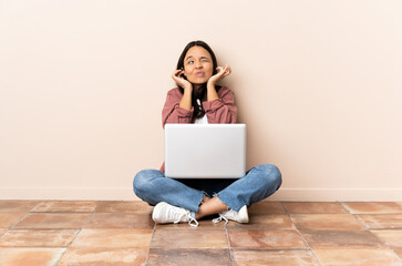 Young mixed race woman with a laptop sitting on the floor frustrated and covering ears