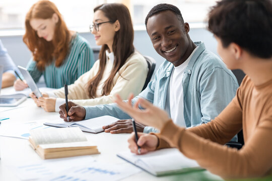 Diverse Group Of Students Sitting At Desk Writing And Talking