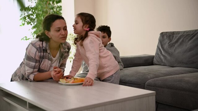 Charming middle aged woman, happy loving mom, enjoying healthy breakfast with her adorable children. A cute girl, daughter whispers in her ear, and laughs, rejoicing in spending time together
