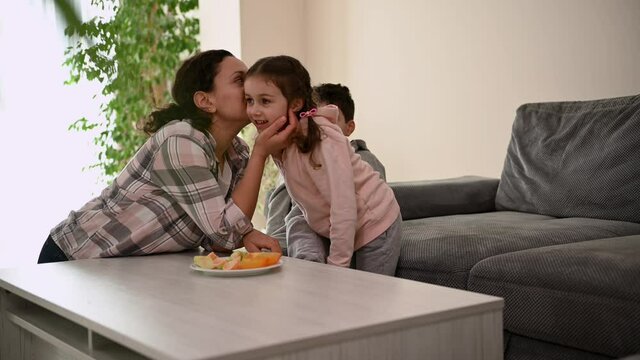 Joyful loving mother whispers a secret in her sweet daughter's ear and everyone together laughs and rejoices sitting at the table at a light healthy snack. Happy mom with her adorable children