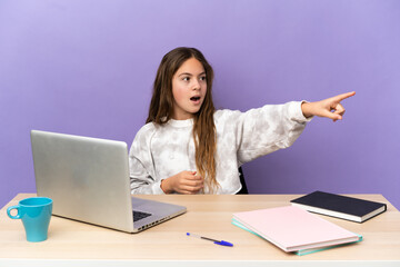 Little student girl in a workplace with a laptop isolated on purple background pointing away