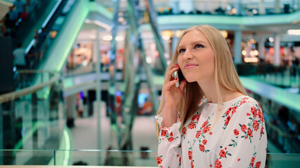 Young woman speaking on phone in mall. Beautiful young woman talking on smartphone and admiring hi-tech interior while standing inside huge mall in Europe.