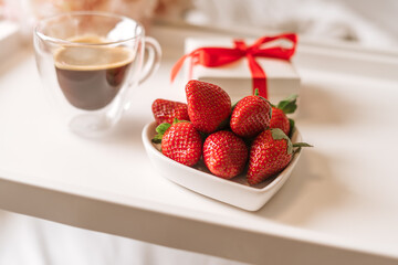 Breakfast for Valentine's Day. Heart shaped white plate with fresh strawberries, cup of coffee and flowers with gift in bed. Still life composition.