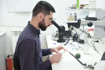 A young man looks through a microscope examining repairs dentures or a jaw in the workshop of a dental technician