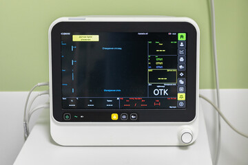 Close up view of a single bedside patient monitor, standing on the table. The screen is on and brightly lit. The wires are plugged in. The monitor is not connected to the patient, but is ready for use