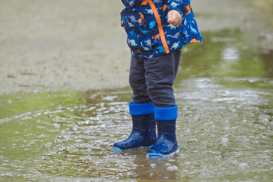Closeup photo of child in waterproof blue rubber boots and raincoat in early spring, child have fun and standing in the puddles after the rain in the city