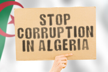 The phrase " Stop Corruption in Algeria " on a banner in men's hand with blurred Algerian flag on the background. Forbidden. Prevent. Wealth. Offence. Corruptness. Economy. Corruptible. Political