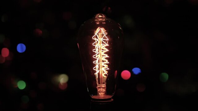 Close Up of vintage incandescent filament bulb switches on and off. Dark blurred background with christmas tree and led lights. Energy expense and electric bills concept.