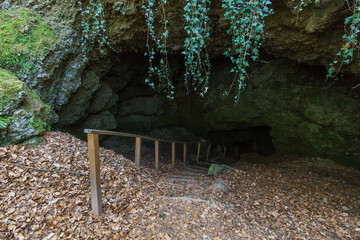 Entrance of a cave in forest with stairs and railing down into the rock, Eishoehlen, Birresborn,...