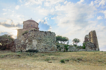 Jvari Monastery - clifftop orthodox monastery dating to circa 590 CE, in Georgia. Blue sky with clouds, ancient stone and brick church walls, dry yellow grass, gren trees