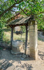 Traditional georgian well in Tbilisi open-air ethnographical museum. Wooden roof, stone pillar, dry grass, green trees and bushes, traditional house on the background