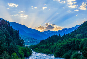 Sunset landscape near the town of Mestia, Svaneti, Georgia. The last rays of sunshine in the blue sky with golden clouds above the blue mountains, green forest trees and white water of the stream