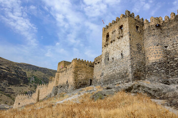 Fototapeta na wymiar Khertvisi fortress in Georgia, Caucasus. Ancient weathered medieval grey and biege stone walls and towers, grey cliff, blue sky with clouds, dry yellow grass