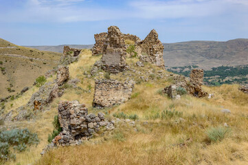 Ruined medieval Tmogvi fortress and Caucasian mountains in southern Georgia. Ancient stone walls and towers, dry yellow grass, green trees, rocky slopes, blue sky with clouds