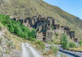 Fototapeta na wymiar The Fortified Village of Shatili, Khevsureti, Georgia. Abandoned towers and houses, brown and grey old stone walls, green trees and dry grass on mountain slopes, blue sky with clouds, road and river