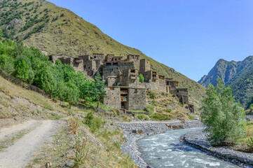 Fototapeta premium The Fortified Village of Shatili, Khevsureti, Georgia. Abandoned towers and houses, brown and grey old stone walls, green trees and dry grass on mountain slopes, blue sky with clouds, road and river